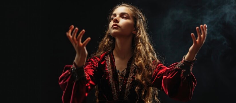 Young witch in costume performs magical gestures with both hands, photographed on a black background.