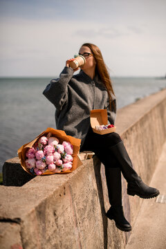 Young woman sits on a concrete slab, drinks coffee and holds a box of macaroons, next to a bouquet