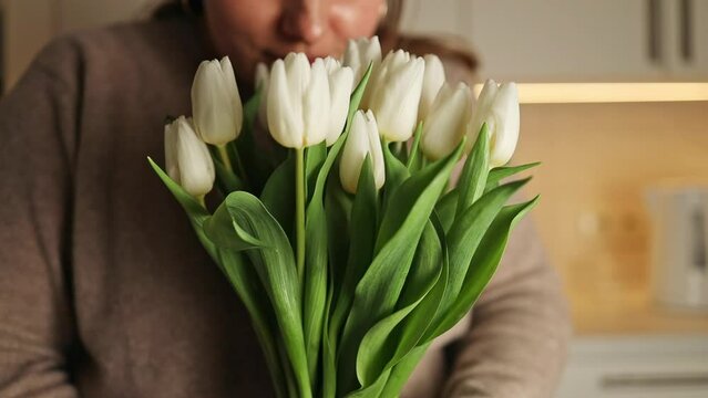 Closeup image of carefree happy 30s woman with tulips flowers for home, close eyes smiling and sniffing beautiful white bouquet. Housewife taking care of coziness in apartment. . High quality FullHD
