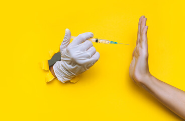 The nurse hand in a white latex glove holds a syringe for vaccination, but collides with a hand in the form of a Stop gesture. Coronavirus anti-vaccination concept.Yellow paper, torn hole, copy space