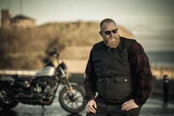 Portrait of a handsome brutal biker with beard, wearing biker clothes and sunglasses. Bike and nature landscape on the background