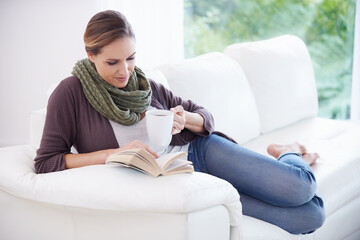 Obraz premium Woman, book and reading with coffee on sofa to relax for story, novel and knowledge in living room at home. Lady, fiction books and break with cup of tea for literature, hobby or comfortable on couch