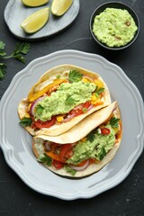 Delicious tacos with guacamole and vegetables served on black table, flat lay