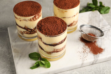 Delicious tiramisu in glasses, mint and sieve with cocoa powder on grey textured table, closeup