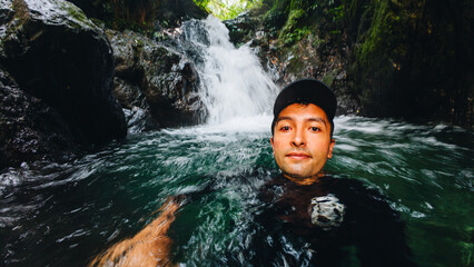 young man swimming in a waterhole of a waterfall in a forest with trees on vacation