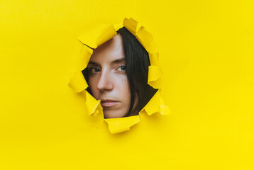 Close-up portrait of a caucasian young woman looking through a hole in yellow paper. An incredulous...