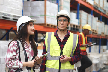 Young Asian female worker wearing safety vest and helmet, holding tablet, discussing with senior...
