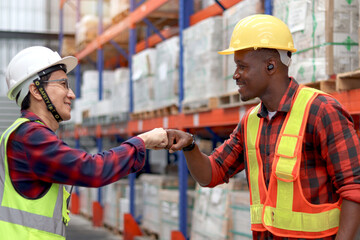 Happy Asian senior supervisor wearing safety vest and helmet, fist bumping hand after complete work with African colleague at cargo logistics warehouse. Worker staffs greet each other with fist bumps.