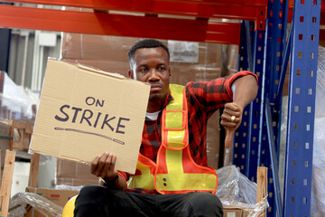 Angry unhappy African worker man wearing safety vest and giving thumb down with strike banner placard sign at cargo logistic warehouse. Striking worker protesting at workplace. - obrazy, fototapety, plakaty