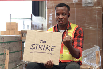 Angry unhappy African worker man wearing safety vest and giving the middle finger with strike banner placard sign at cargo logistic warehouse. Striking worker protesting at workplace.