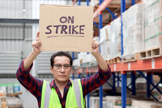 Angry unhappy Asian senior worker wearing safety vest, holding and raising sign on strike banner placard at cargo logistic warehouse. Striking worker protesting at workplace.
