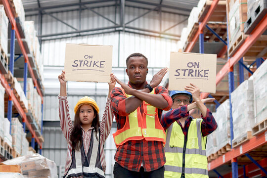 Angry unhappy African worker with colleagues, Asian senior and woman staffs wear safety vest and helmet, hold sign on strike banner at cargo logistic warehouse. Striking worker protesting at workplace