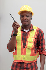 Portrait of young African worker man wearing safety vest and helmet, holding walkie talkie on gray walls. Engineer talking to a colleague with walkie talkie at logistic shipping cargo warehouse.