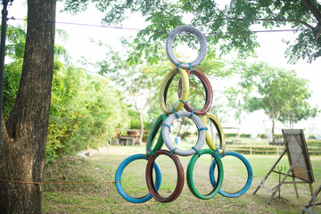 A creative obstacle course feature made of colorful, painted, recycled tires stacked in a pyramid hanging from a tree in a sunny park. A fun outdoor activity for fitness and team building.