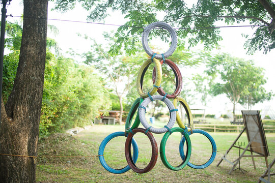 A creative obstacle course feature made of colorful, painted, recycled tires stacked in a pyramid hanging from a tree in a sunny park. A fun outdoor activity for fitness and team building.