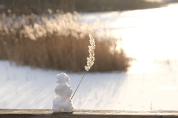 Little Snowman standing on bridge in front of a frozen lake waving with a reed in backlight of...