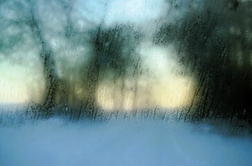 Abstraktes Motiv mit feuchter Eis bedeckter Glasscheibe vor schneewiese mit Bäumen vor weiß-rotem Himmel bei Schnee,  Kälte, Eis und Schnee bei Sonnenuntergang am Nachmittag im Winter