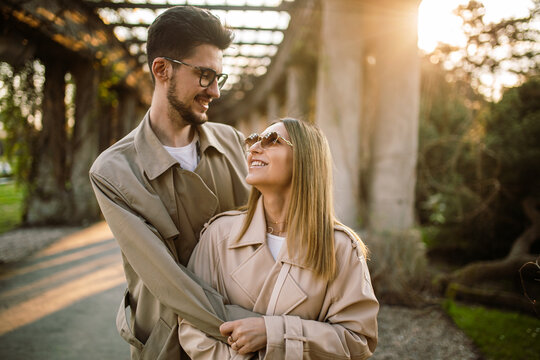 Portrait of a girl and a guy posing against the backdrop of an arch in the garden at sunset in spring. Family photo of a stylish couple in beige trench coats.