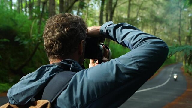 Active adventure middle age man travel photographer with backpack shooting by camera beautiful forest landscape with asphalt road. Back view.