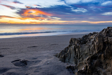 Breathtaking sunset over a peaceful beach, with footprints in the sand and rocky formations. Perfect for travel, nature, and relaxation themes in wall art, home décor, or marketing materials.