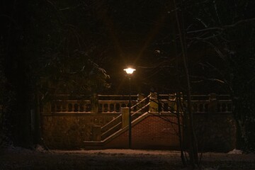 Zimowy wieczór w parku. Samotna, elektryczna lampa oświetlająca kamienne schody o konstrukcji murowanej z czerwonej cegły. Ziemię pokrywa warstwa białego śniegu. - obrazy, fototapety, plakaty