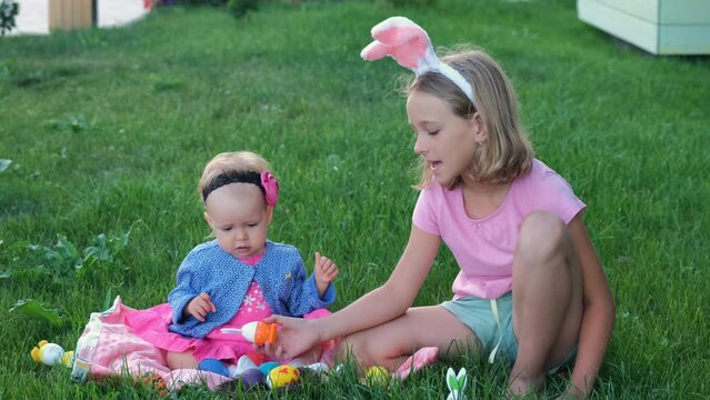 Little girl in bunny ears with adorable baby sister playing with colorful eggs on the grass, slow motion. Easter hunt concept