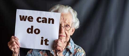 Old Caucasian woman holding a paper with text "we can do it" indoors, in closeup.