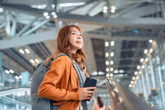 Happy asian tourist woman traveller with backpack using mobile phone while stand on moving walkway in airport terminal, Tourist journey trip concept.