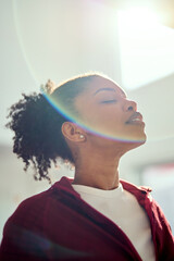 Young healthy calm serene African American woman wearing sportswear standing with eyes closed lit with sunlight doing yoga exercises breathing fresh air. Close up. Mental balance concept. Vertical