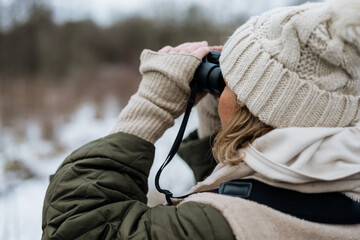 Woman dressed in winter clothes with gloves and knitted beanie looking for birds and animals through the binoculars in nature - obrazy, fototapety, plakaty