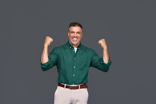 Happy confident middle aged business man entrepreneur, excited older professional businessman winner raising arms celebrating victory and success standing isolated on gray background.