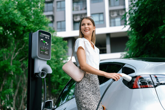 Young woman travel with EV electric car charging in green sustainable city outdoor garden in summer. Urban sustainability lifestyle by green clean rechargeable energy of electric BEV vehicle innards