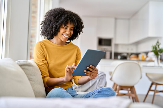 Happy young African American woman sitting on couch at home using digital tablet. Smiling relaxed lady relaxing on sofa looking at tab technology device holding pad computer in hands in living room. - Powered by Adobe