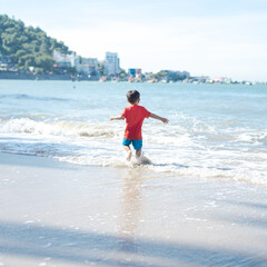 Toddler boy Asian kid running playing with waves along brown sandy beach shoreline with Vung Tau city buildings hotels skylines background mountain range, Vietnam travel destination