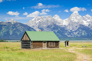 Fototapeta na wymiar Historic wood log building in the valley meadow in Grand Teton National Park with the snow-capped Teton Mountain Range and blue sky with clouds in the background.