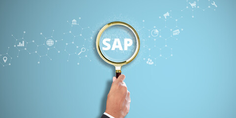 SAP: Businessman Hand holding a magnifying glass with SAP icon on Light Blue background. Enterprise Resource Planning, Business Software Solutions, Digital Transformation.