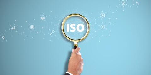 ISO: Businessman Hand holding a magnifying glass with ISO icon on Light Blue background. International Standards Organization, Quality Assurance, Compliance Excellence.