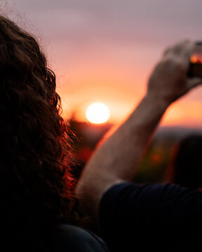 Portrait Sunlight shines through the hair of a person viewing a beautiful sunset while another person is taking a picture with their phone. The horizon beautiful purple and orange with the sun visible