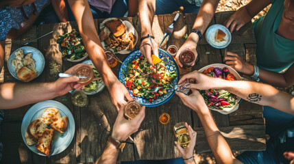 Top view of a group of people sitting around a rustic wooden dining table, toasting with their glasses raised amidst a spread of various dishes - obrazy, fototapety, plakaty