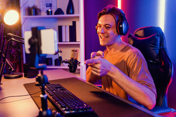 Fototapeta na wymiar Host channel of gaming smart streamer playing online game, wearing headphone with viewers live steaming on media social online by smartphone talking with team player at neon lighting room. Pecuniary.
