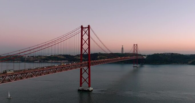 Aerial drone shot towards the 25th April Bridge, Ponte 25 de Abril, and Cristo Rei in Lisbon, Portugal, Europe. Pink and orange sky after sunset, end of autumn, start of winter. Shot in ProRes 422 HQ