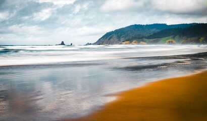 long exposure of waves washing over sand along the Oregon coast with passing rain clouds above the mountainous landscape. 