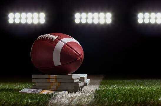 Low angle view of a football sitting on a pile of money on a grass field below stadium lights
