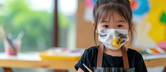 A young Asian girl, wearing a face mask, is painting with watercolors. The cute child, wearing a black apron, is 3 years old.