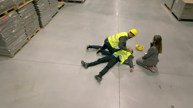 Colleagues giving first aid to unconscious worker after accident at work in warehouse, factory. Calling the ambulance. Concept of occupational safety and health in workplace.
