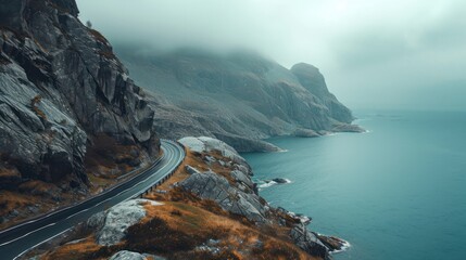  a winding road on the side of a mountain with a body of water in the foreground and a cliff on the other side of the road on a foggy day. - obrazy, fototapety, plakaty