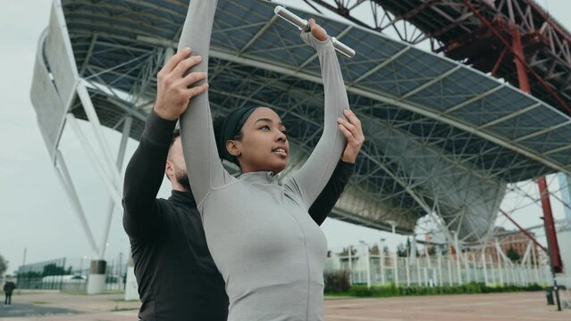 Middle Eastern male trainer instructing Black woman in fitness attire on performing a shoulder press with dumbbells in an urban environment.