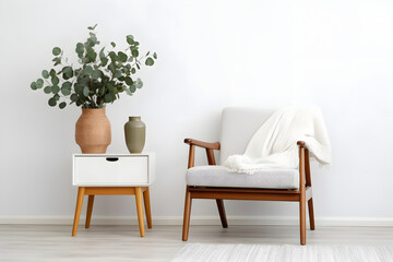 Vase with green eucalyptus branches on end table and comfortable armchair near white wall