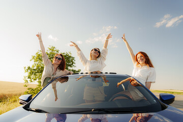 Three friends pop out of a car sunroof, arms raised in joy, embracing the freedom of a summer road trip in the countryside © arthurhidden