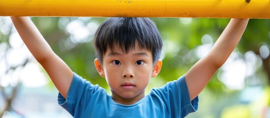 Asian boy exercises outdoors by hanging from a yellow bar.